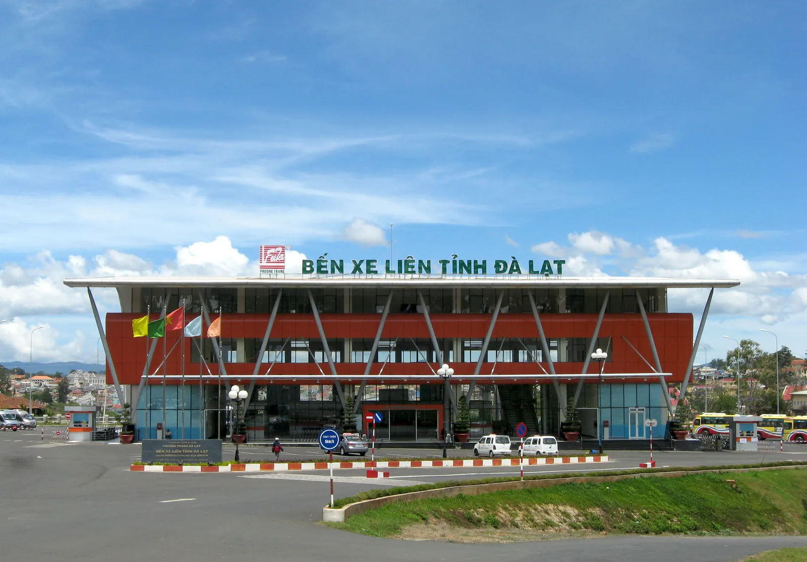 Mặt trước Bến xe liên tỉnh Đà Lạt với dòng chữ tên bến trên mái, mặt tiền màu cam và kính, hàng cột thép trắng xếp chéo, vài xe con đỗ trước sảnh và xe khách đỗ ở hai bên dưới trời xanh