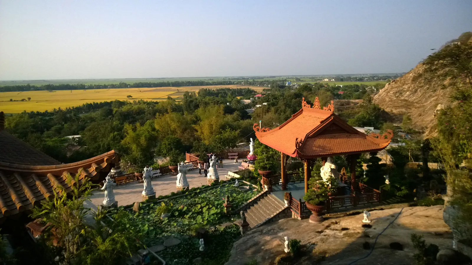 Sân chùa bậc thang trên sườn Núi Sam ở Châu Đốc: nhà mát mái ngói đỏ, hàng tượng trắng và hồ sen, bậc thang đá nối hai tầng sân, vách núi bên phải và đồng lúa chín vàng của đồng bằng trải rộng phía sau